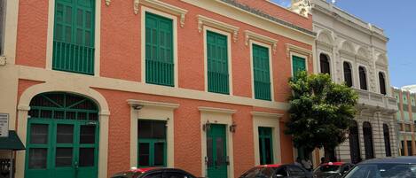 A building with green shutters and a white facade, parked cars, and a clear blue sky.