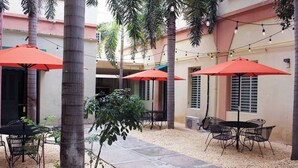 An outdoor patio area with tables and chairs under red umbrellas, surrounded by palm trees and a building with beige walls and shutters. - Gladiolas 1104 (Ponce)