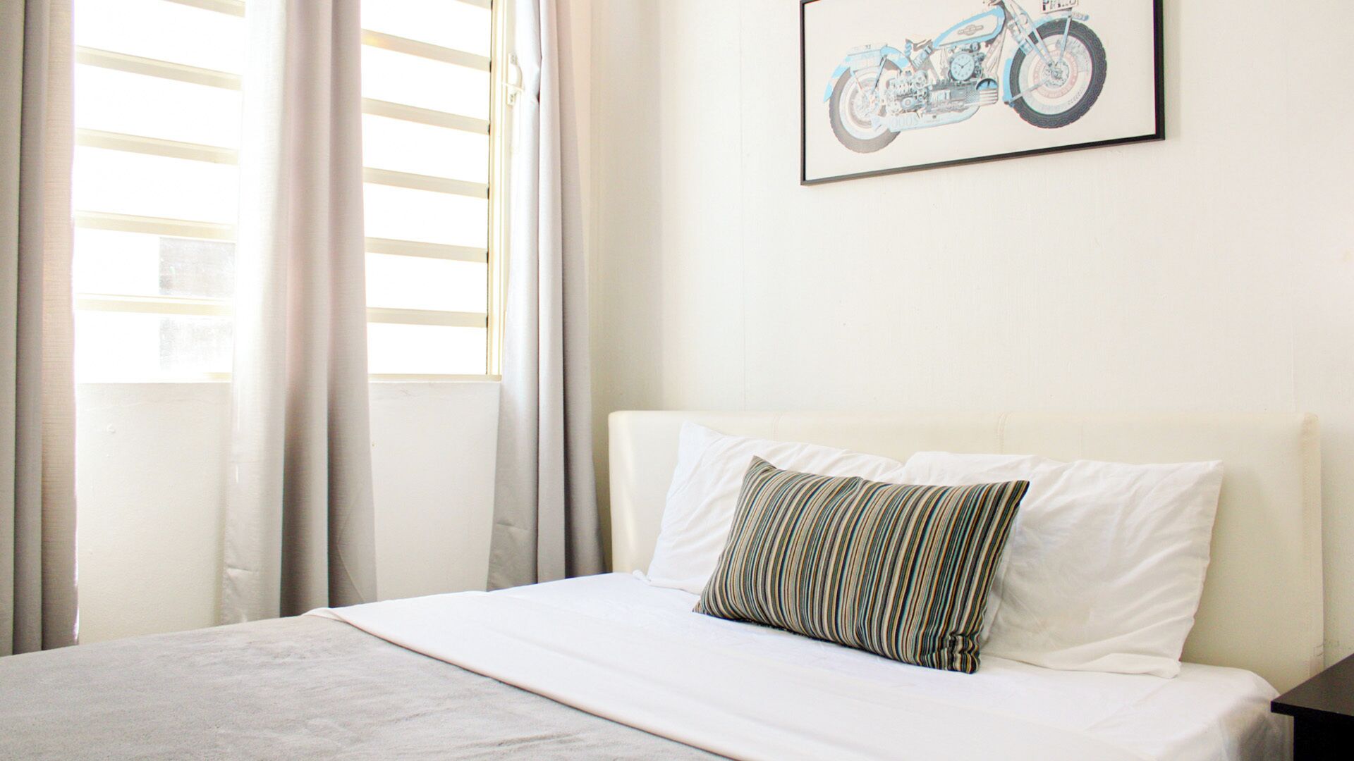 A neatly made bed with white linens and a striped pillow, a framed motorcycle artwork on the wall, and a window with curtains.