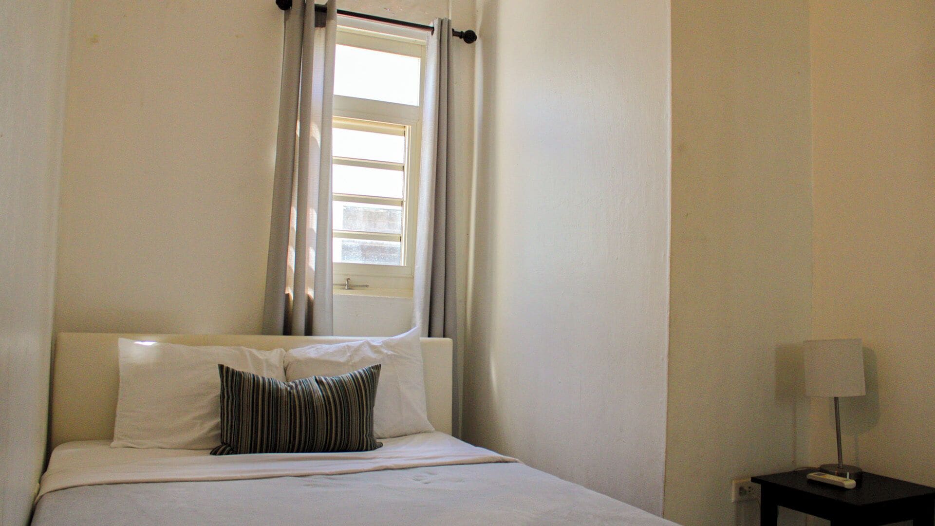 A single bed with white bedding and a striped pillow, a window with curtains, and a small bedside table with a lamp.