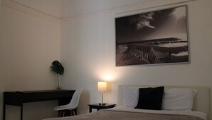 Une chambre d’hôtel comprenant un lit, un bureau, une chaise, une table de chevet, une lampe et un grand tableau encadré représentant une scène de plage.