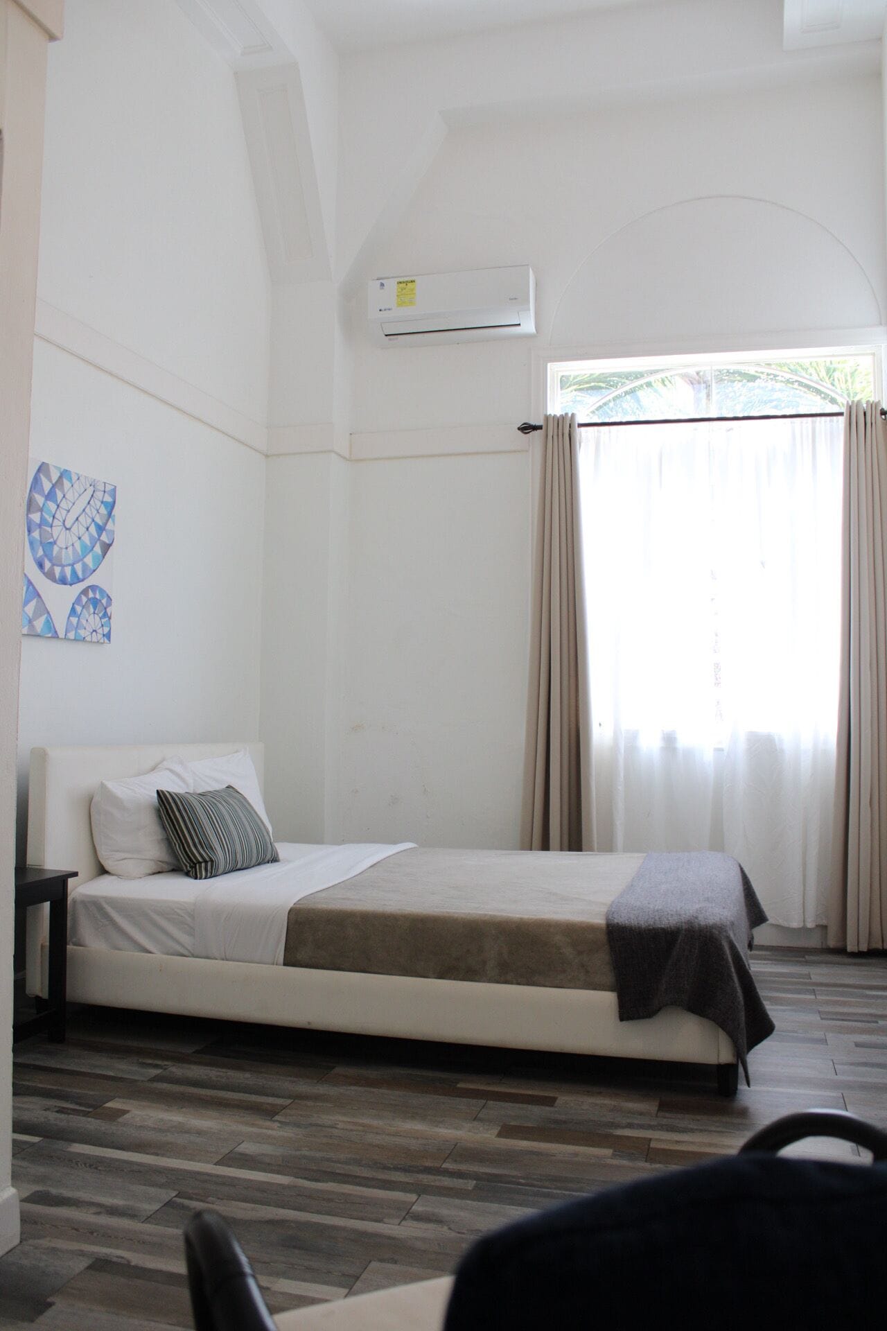 A single-bed room with a wooden floor, a bed with a gray blanket, a pillow, a nightstand, a wall-mounted air conditioner, and a window with curtains.