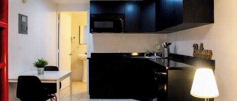 A modern kitchen with black cabinets, a microwave, and a sink. There's a dining area with a table and chairs, and a lamp on the right.