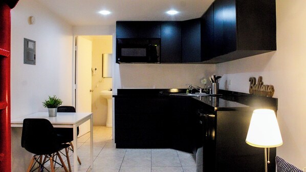Una cocina moderna con gabinetes negros, un microondas y un lavamanos. Hay un comedor con una mesa y sillas, y una lámpara a la derecha.