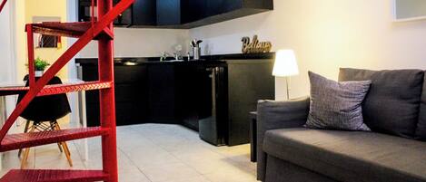 A modern living room with a grey sofa, a red staircase, a kitchen with black cabinets, and a small dining area.