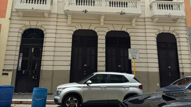 Un edificio in stile architettonico classico, caratterizzato da un ingresso centrale con una grande porta nera, affiancato da due finestre più piccole con persiane. Presenta tre balconi con ringhiere bianche e una facciata chiara con elementi decorativi.