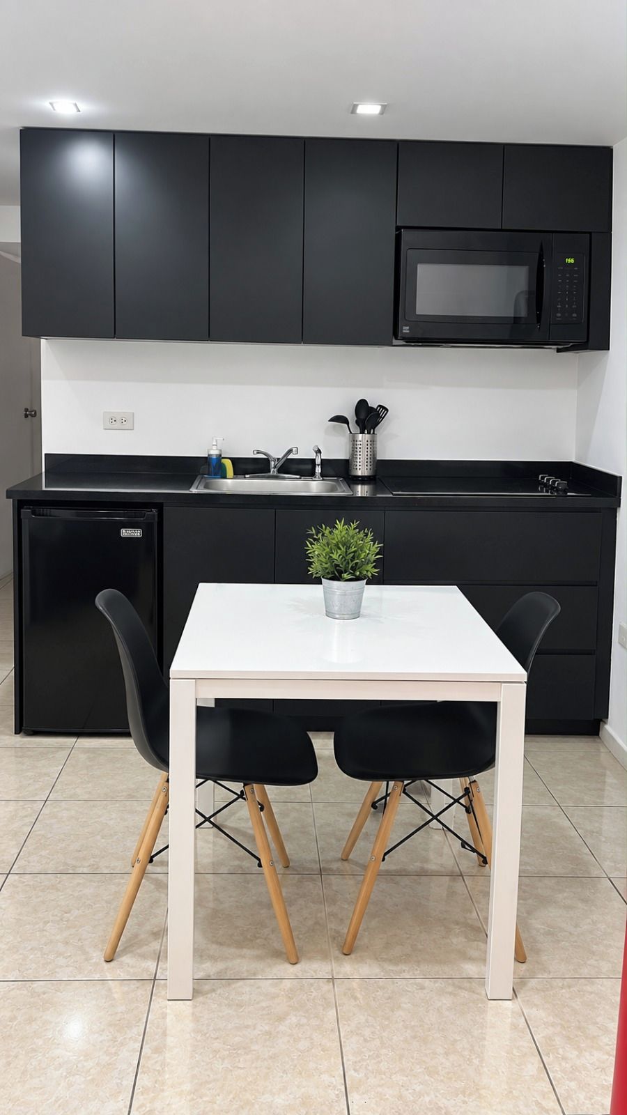 A modern kitchen with black cabinets, a white countertop, a black refrigerator, a microwave, and a small potted plant on the table.