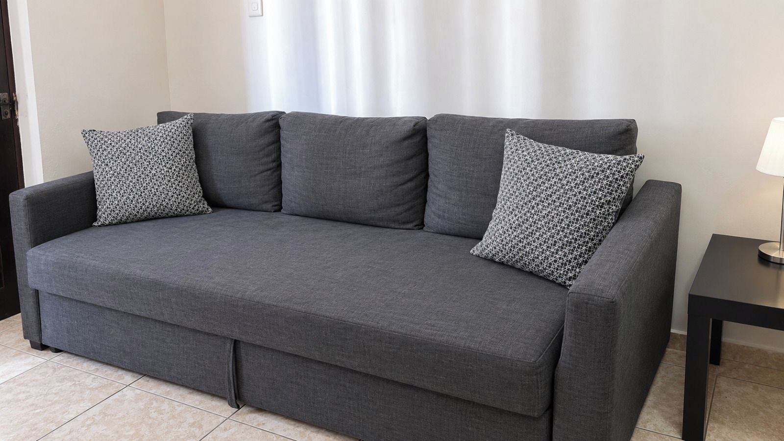 A grey sofa with patterned cushions in a room with a tiled floor and a small table with a lamp.