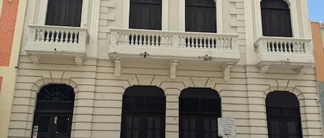 A building with a classic architectural style, featuring a central entrance with a large black door, flanked by two smaller windows with shutters. There are three balconies with white railings, and the building has a light-colored facade with decorative elements.