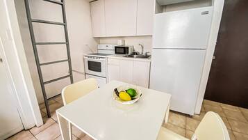A compact kitchen with white cabinets, a microwave, stove, and refrigerator. There is a table with a bowl of fruit and two chairs.