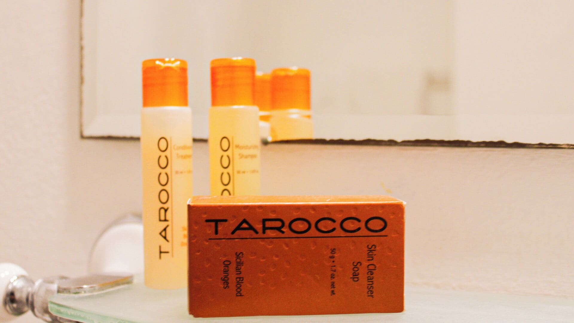 Un baño con un espejo, un lavamanos y tres botellas de productos para el cuidado de la piel etiquetados 'Tarocco'.