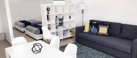 Un salon moderne avec un canapé gris, des chaises blanches, une table à manger blanche et une bibliothèque.