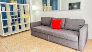 Un salon avec un canapé gris, un coussin rouge, une étagère blanche avec des rangements bleus et jaunes, une lampe sur pied et un miroir au mur.