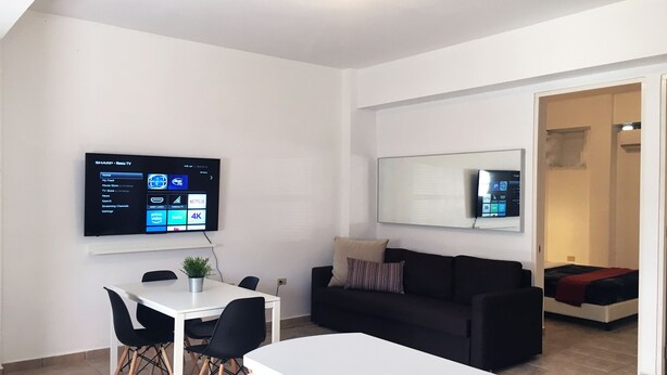 A modern living room with a flat-screen TV mounted on the wall, a black sofa, a white dining table with black chairs, and a small potted plant. - Boulevard 106 (Mayagüez)