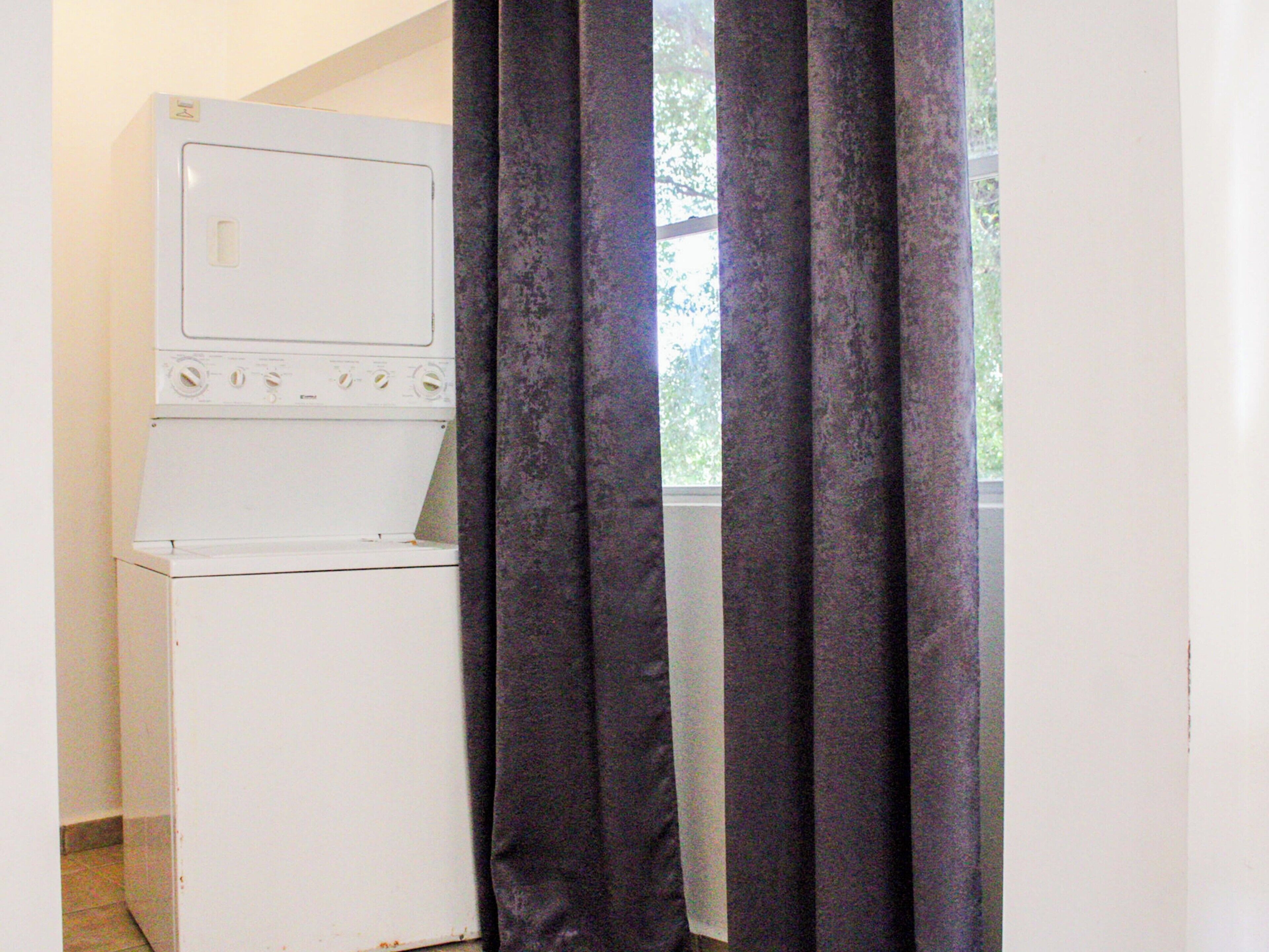 A laundry room with a white washer and dryer, a window with curtains, and a door leading to another room.
