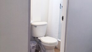 Une petite salle de bain blanche, équipée de toilettes, d’une douche et d’une poubelle.