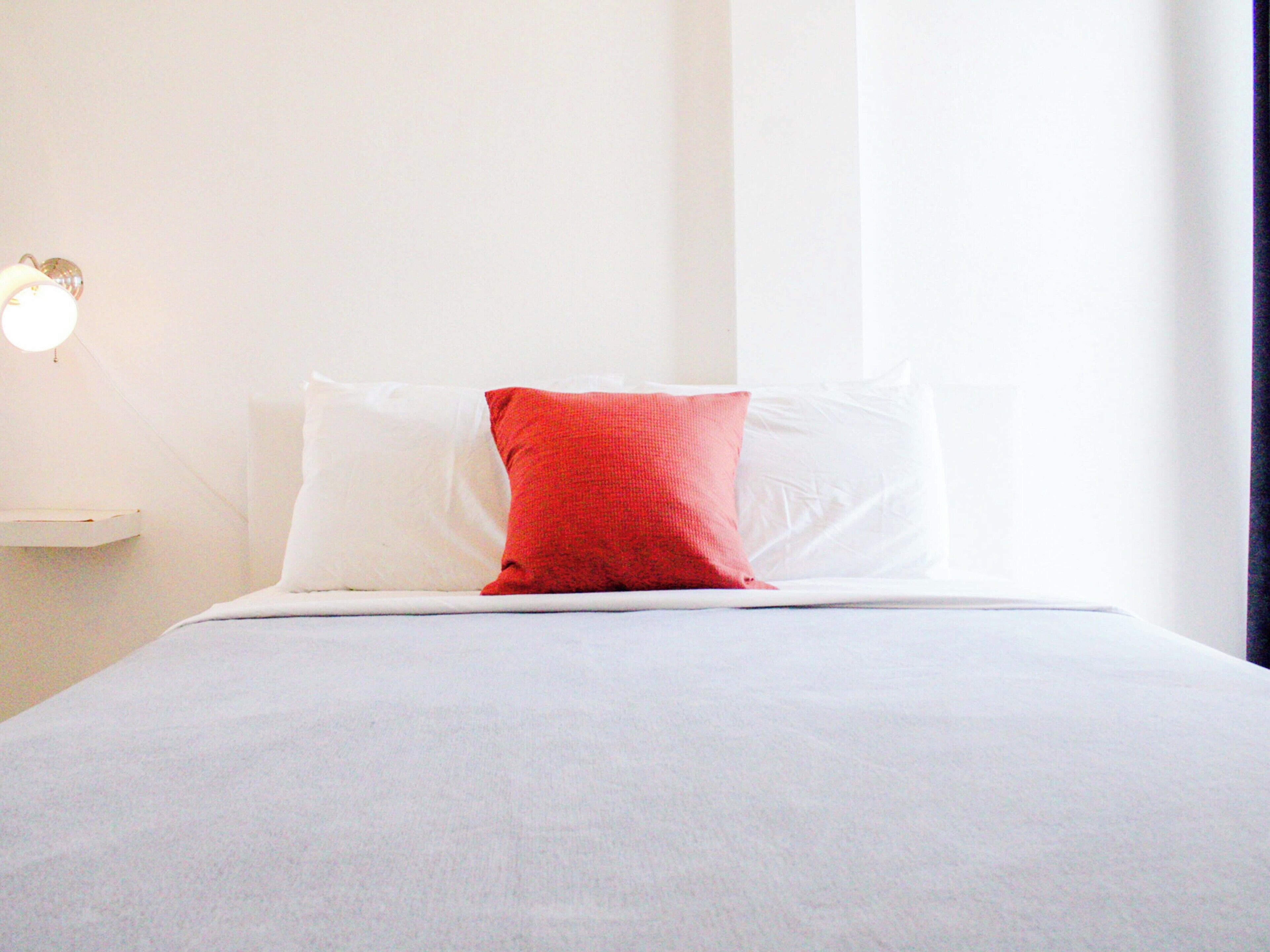 Uma cama com almofadas brancas e vermelhas, uma parede branca e uma mesa de cabeceira com abajur.