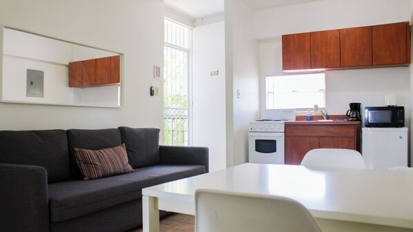 Un espacio habitable pequeño con un sofá gris, una mesa blanca y una kitchenette que incluye estufa, microondas y armarios.