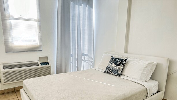 A neatly made bed with a white cover, a patterned pillow, and a white pillow. There is a window with blinds, a radiator, and a curtain. - Boulevard 204 (Mayagüez)