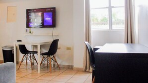 A room with a TV mounted on the wall displaying the Roku interface, a white desk with two chairs, a dining table with chairs, and a window with curtains. - Boulevard 207 (Mayagüez)