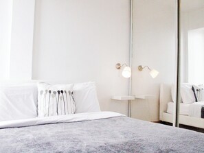 Un lit bien fait, avec du linge de lit blanc et une couverture grise à texture intéressante, une table de chevet avec une lampe et un grand miroir accroché au mur.