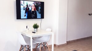 Ett modernt vardagsrum med en platt-TV som är monterad på väggen, ett vitt matbord med två stolar, en liten krukväxt och en grå matta på golvet.