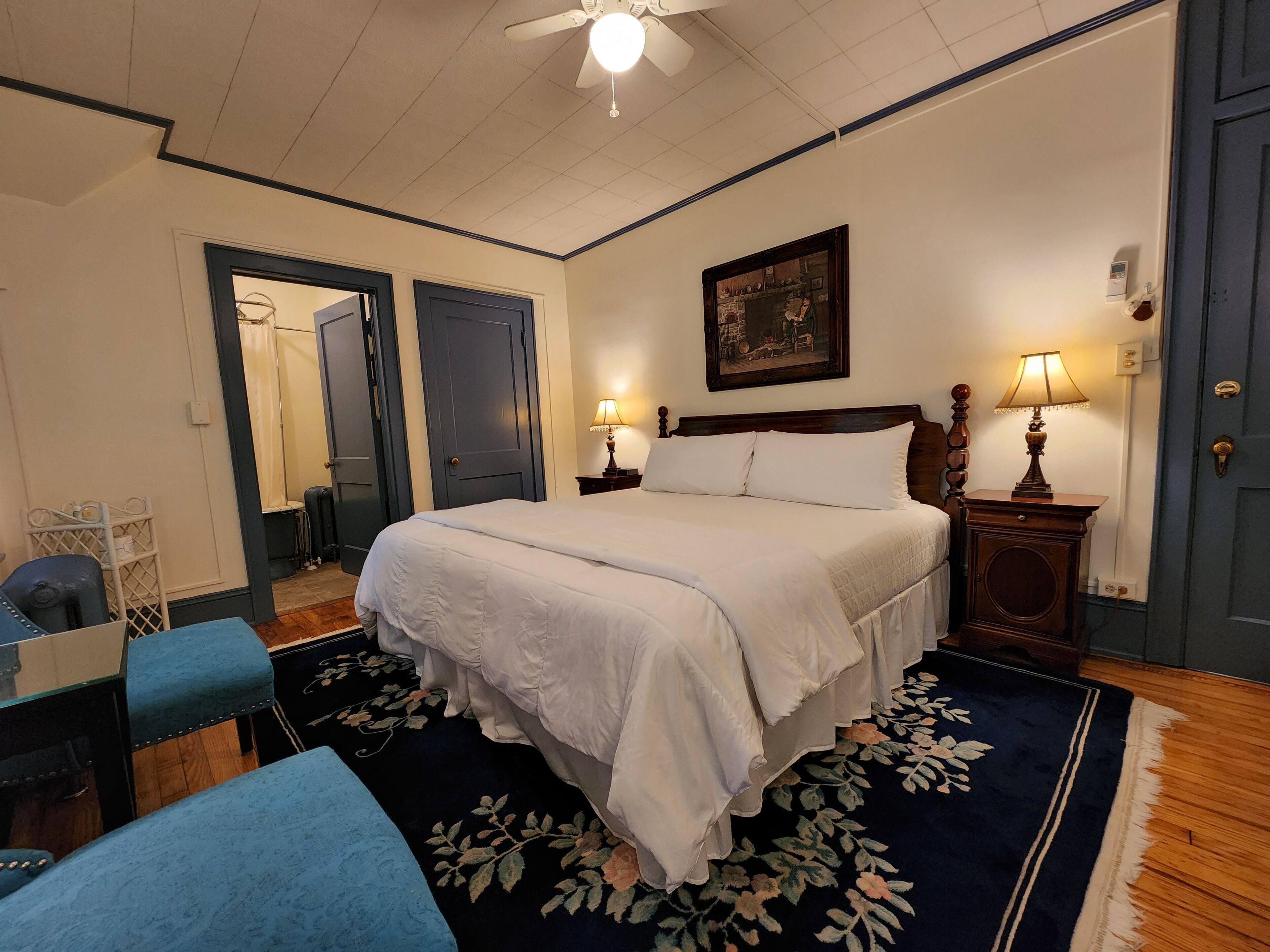 A bedroom with a large bed, two bedside lamps, a ceiling fan, a painting on the wall, and a door with a mirror.