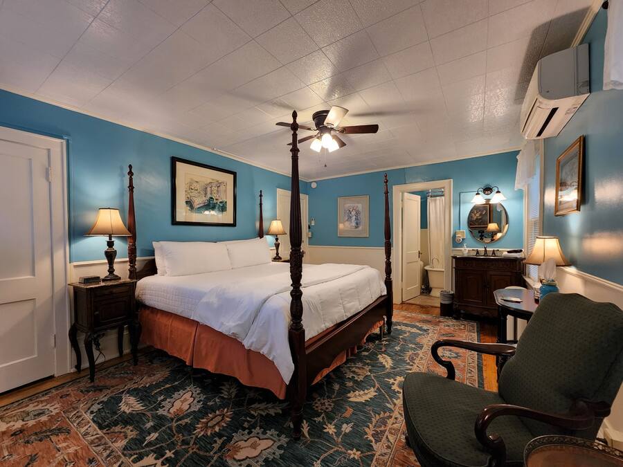 Un lit à baldaquin, un fauteuil, une table de chevet avec une lampe, une salle de bain avec un rideau de douche et un climatiseur fixé au mur.