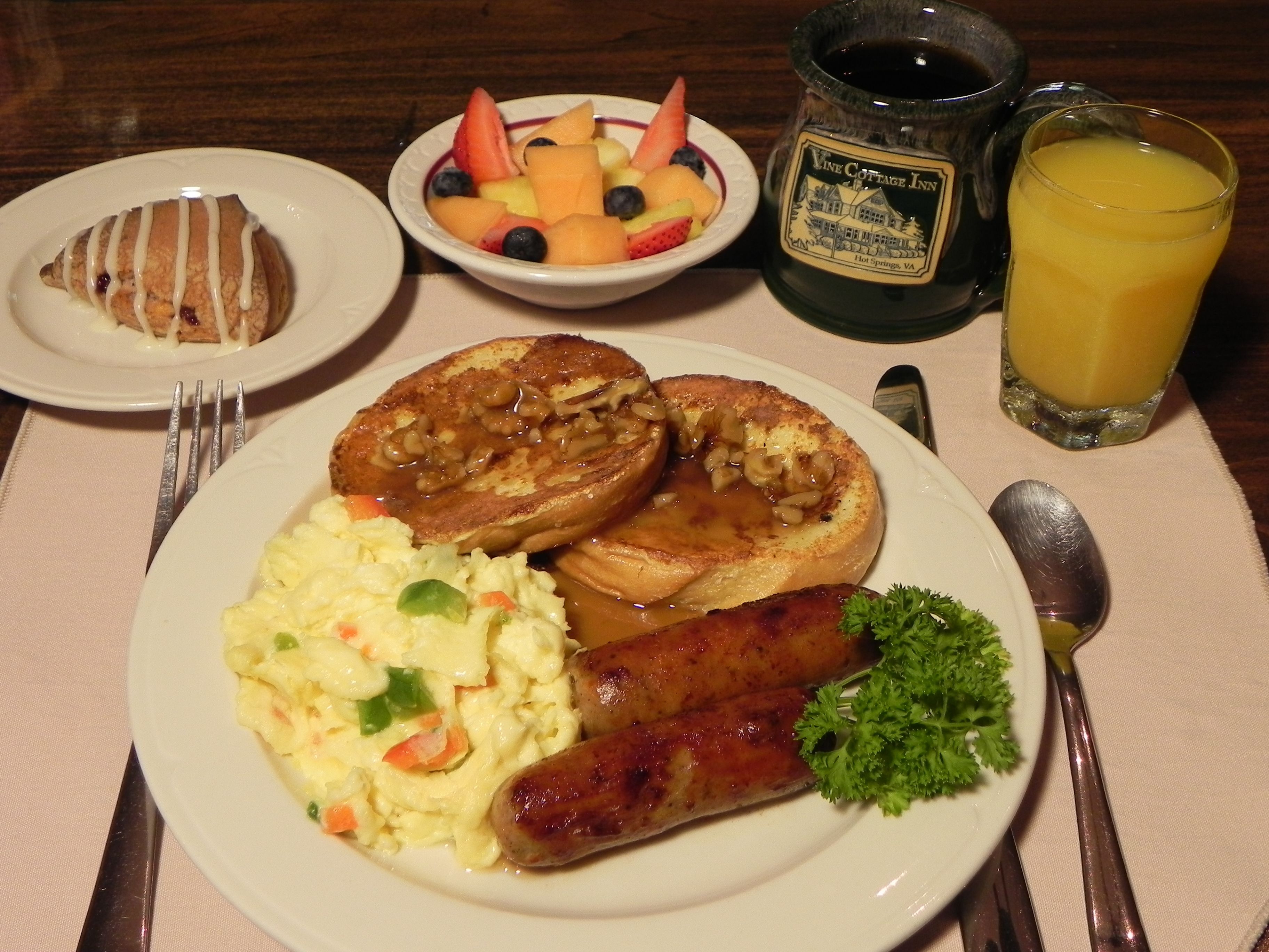 Un plato de desayuno con salchicha, huevos revueltos, tostadas y un bol de fruta.