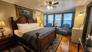 A bedroom with a large bed, a ceiling fan, a window with blinds, a chair, a nightstand, and a lamp. - Vine Cottage Inn (Hot Springs)