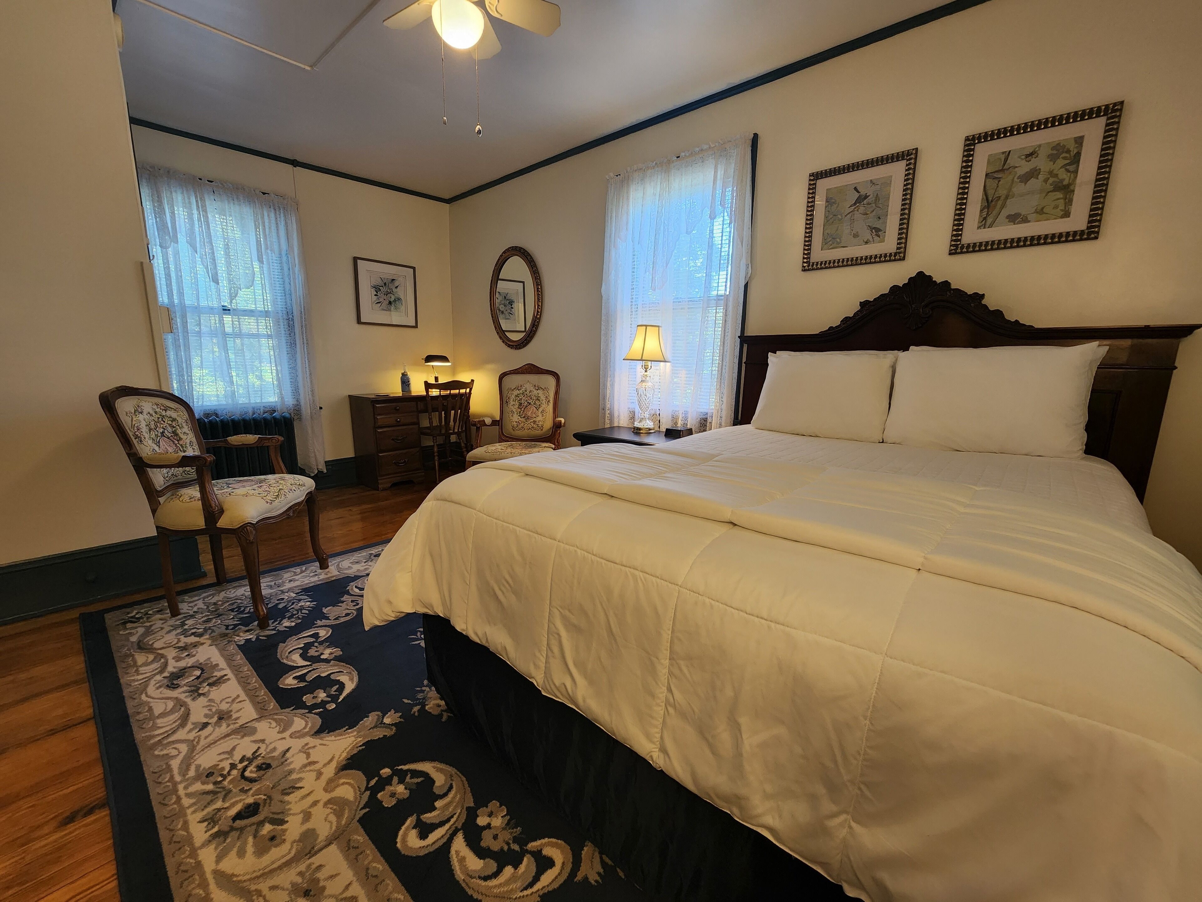 A bedroom with a large bed, two chairs, a dresser, and a desk. There are two windows with curtains, a ceiling fan, and framed pictures on the wall.