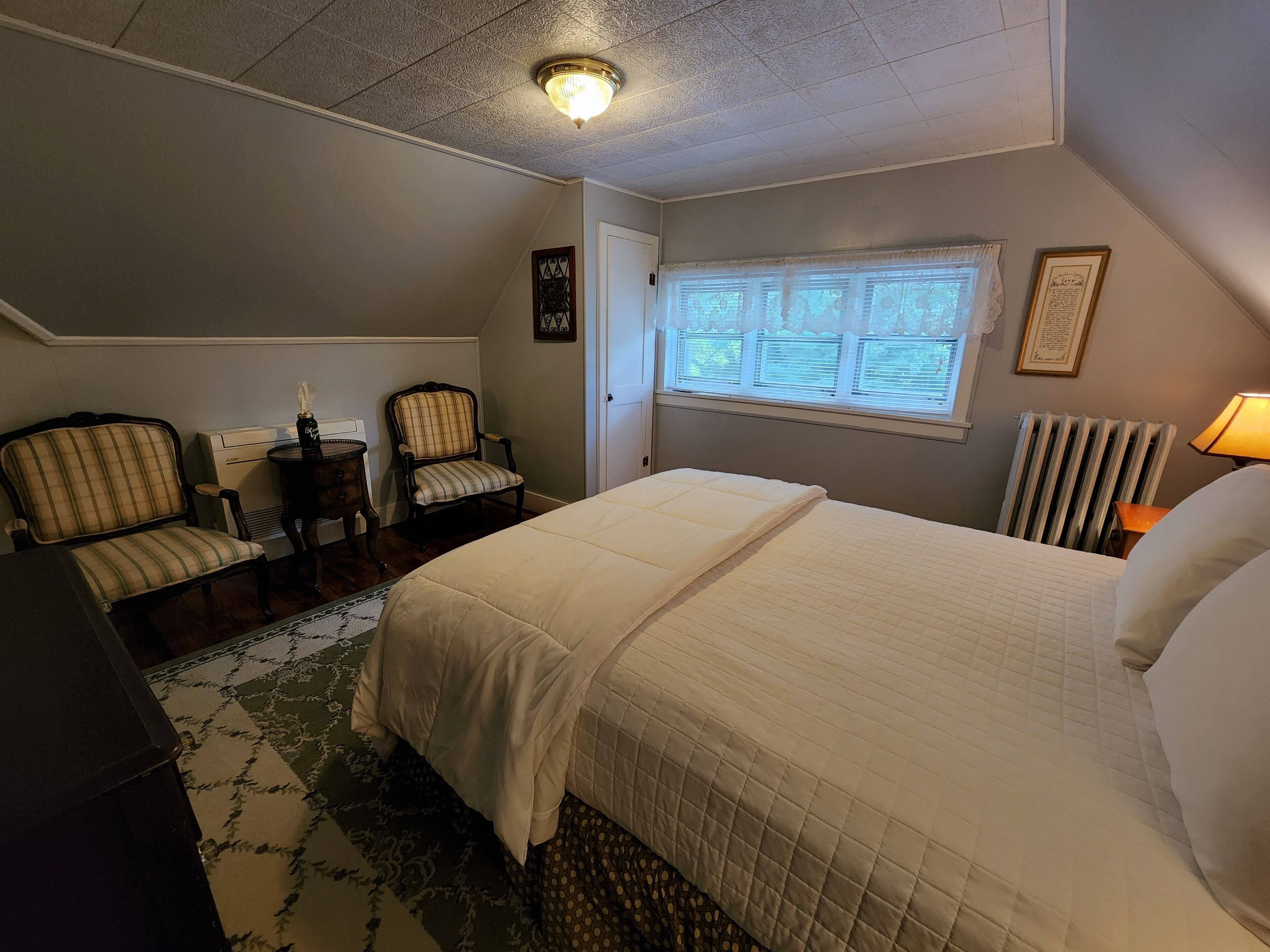A bedroom with a large bed, two chairs, a small table, a radiator, and a window with blinds.