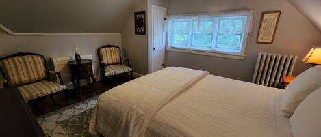A bedroom with a large bed, two chairs, a small table, a radiator, and a window with blinds.