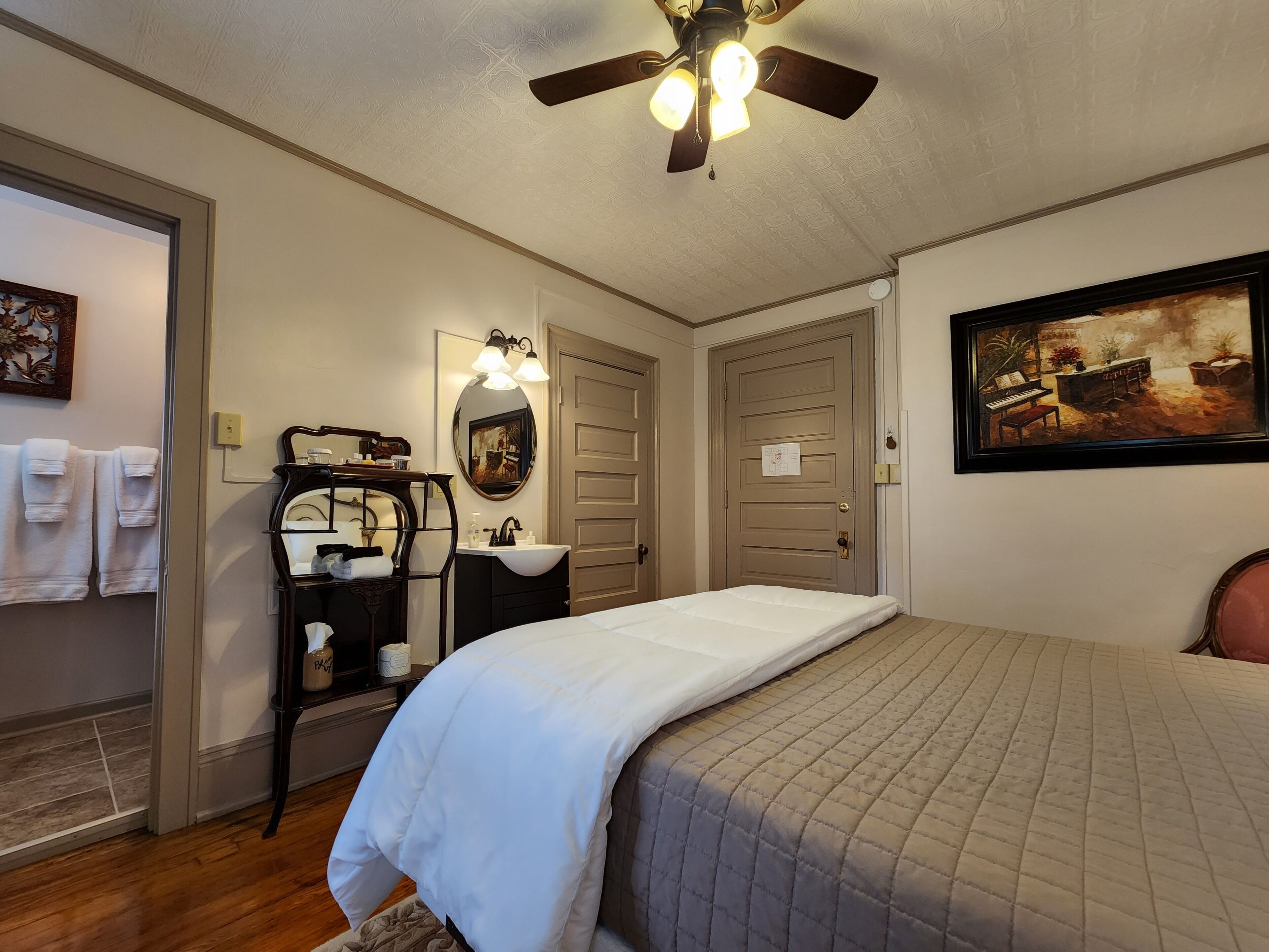A hotel room with a bed, a vanity with a mirror, a ceiling fan, and a painting on the wall.