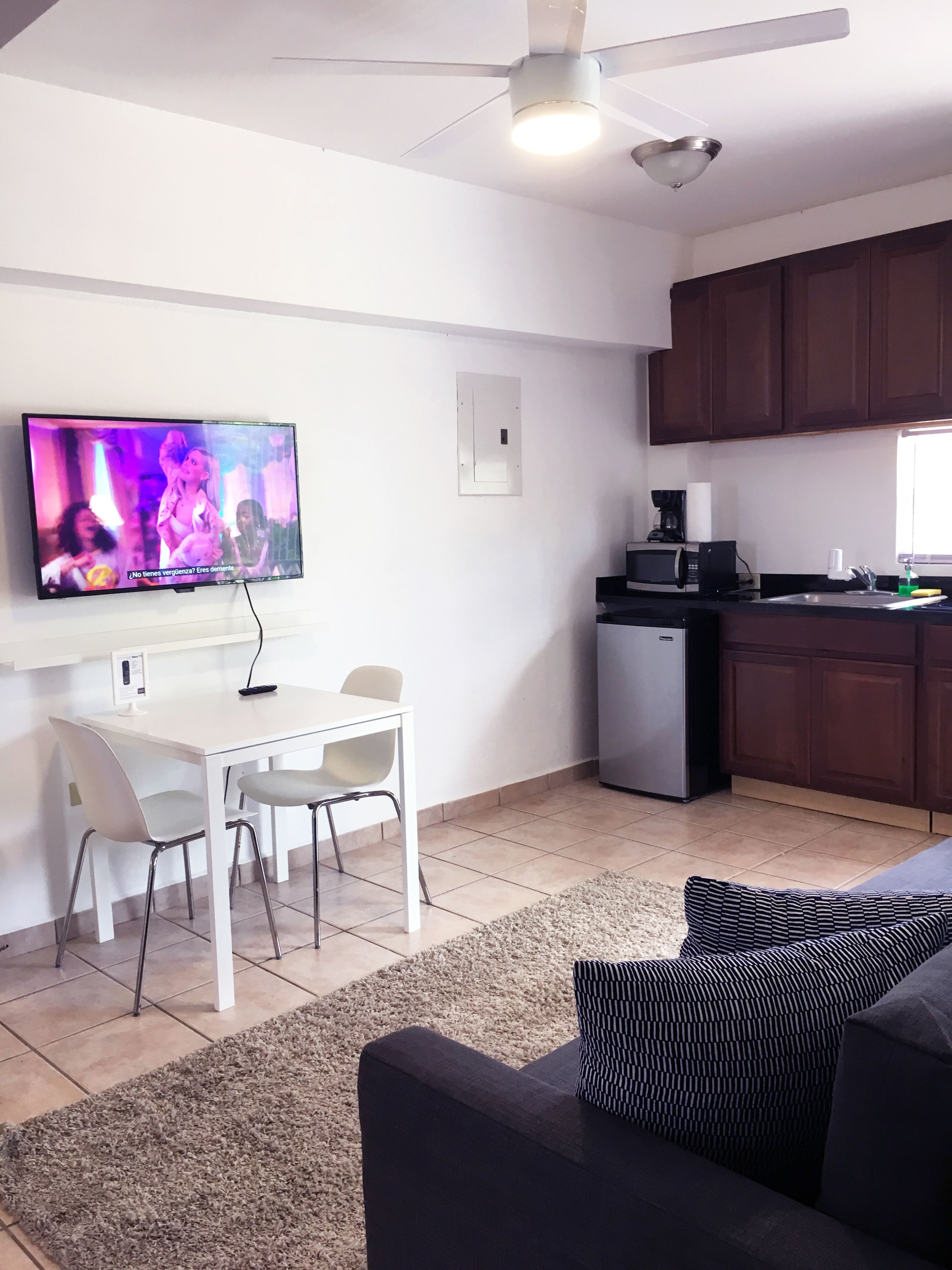Una sala de estar moderna con un televisor de pantalla plana, un comedor con una mesa y sillas blancas, una cocina con armarios de madera oscura y un ventilador de techo.
