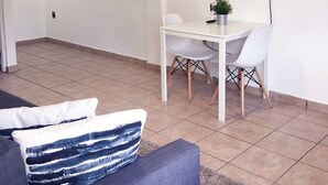 Un salon moderne avec un téléviseur à écran plat fixé au mur, une table à manger blanche avec deux chaises, un canapé gris avec des coussins à motifs et une petite plante en pot posée sur la table.