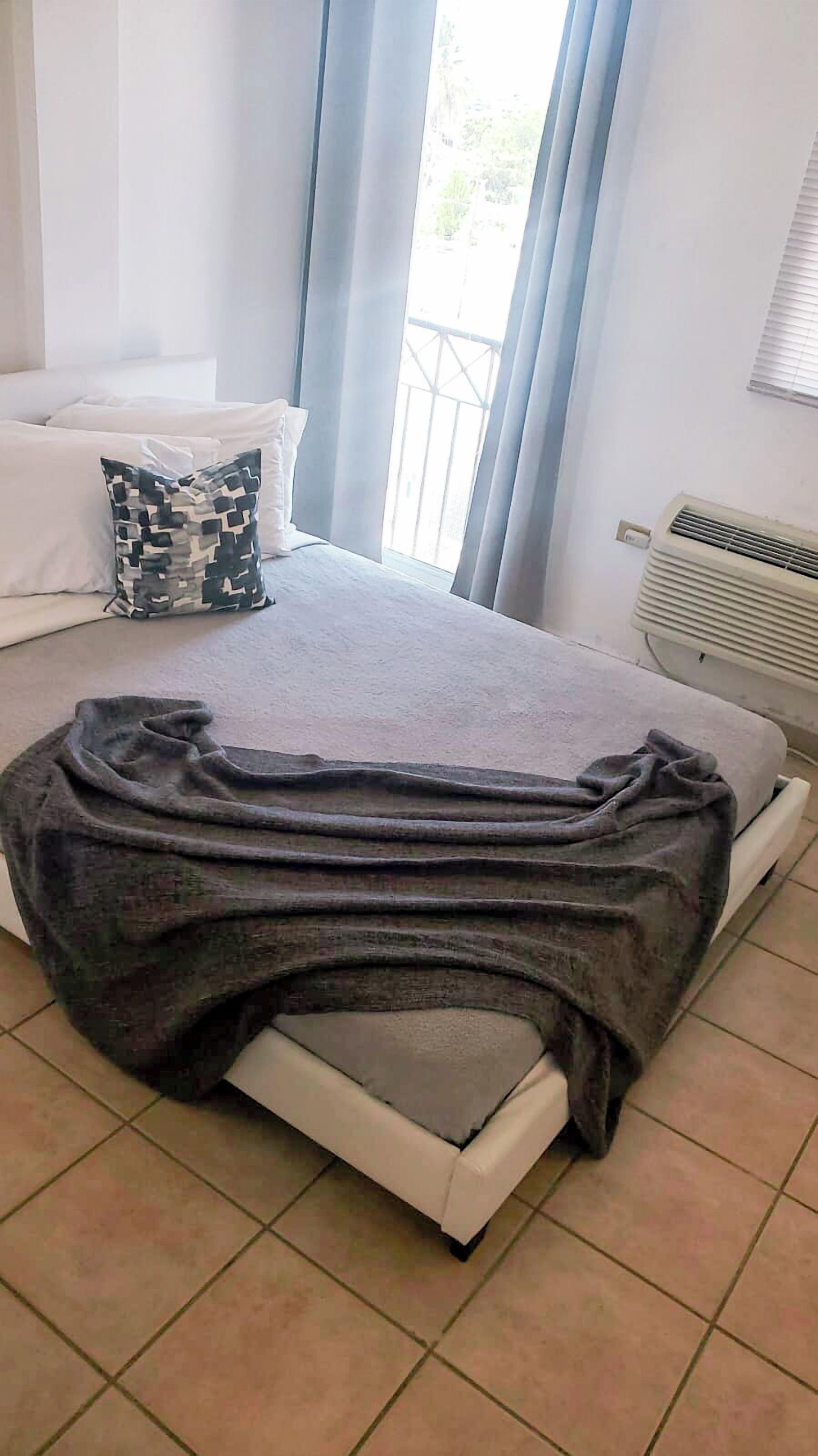 Un dormitorio con una cama, un almohadón, una ventana con cortinas y un equipo de aire acondicionado.