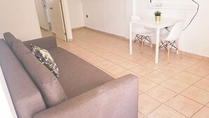 Un salon avec un canapé gris, une petite table accompagnée de deux chaises et une plante en pot.