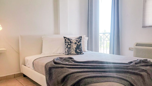 Une chambre à coucher avec un lit, des oreillers blancs, un coussin à motifs, une couverture, un radiateur et une fenêtre avec des rideaux.