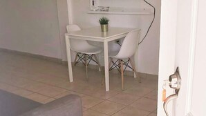 Une pièce avec un canapé gris, une table blanche et deux chaises, une petite plante posée sur la table et un téléviseur fixé au mur.
