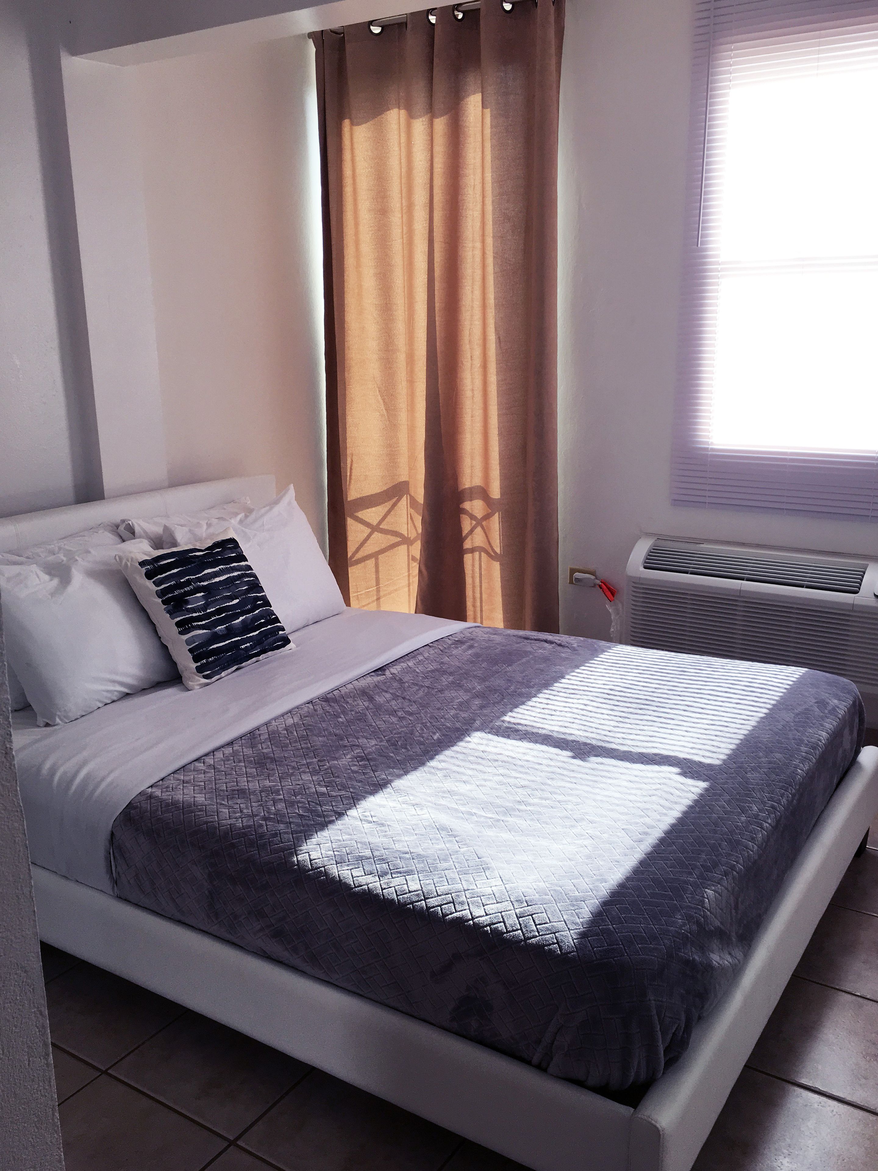 A neatly made bed with a quilted cover, a pillow with a geometric pattern, and a window with blinds.