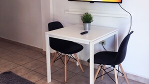 Une petite pièce moderne avec une table blanche et deux chaises noires, une plante en pot et un téléviseur à écran plat fixé au mur.