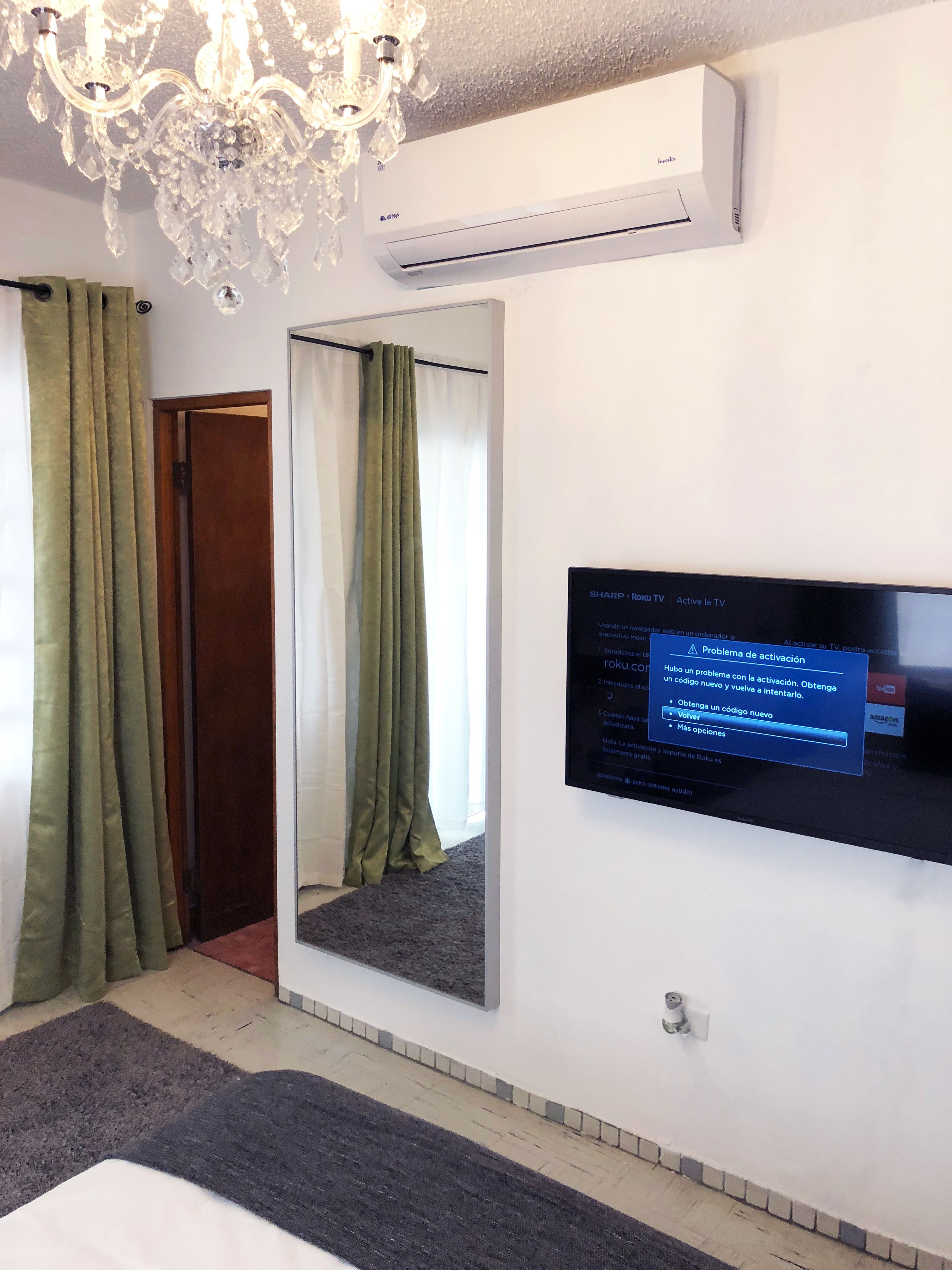 A hotel room with a flat-screen TV mounted on the wall, a large mirror, a chandelier, and green curtains.