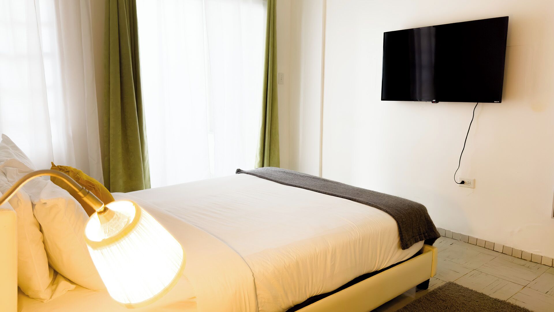 Habitación de hotel con cama, lámpara de noche, televisor de pantalla plana en la pared y ventana con cortinas.