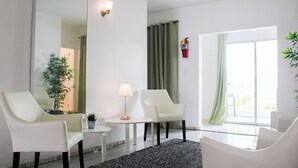 A modern living room with two white armchairs, a small round table with a lamp, a potted plant, and a view of a green curtain and a fire extinguisher on the wall. - Aibonito Hotel 202 (Aibonito)