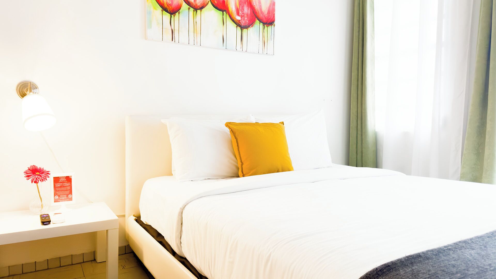 A hotel room with a bed, white bedding, a nightstand with a lamp, a red flower, and a painting of red tulips on the wall.