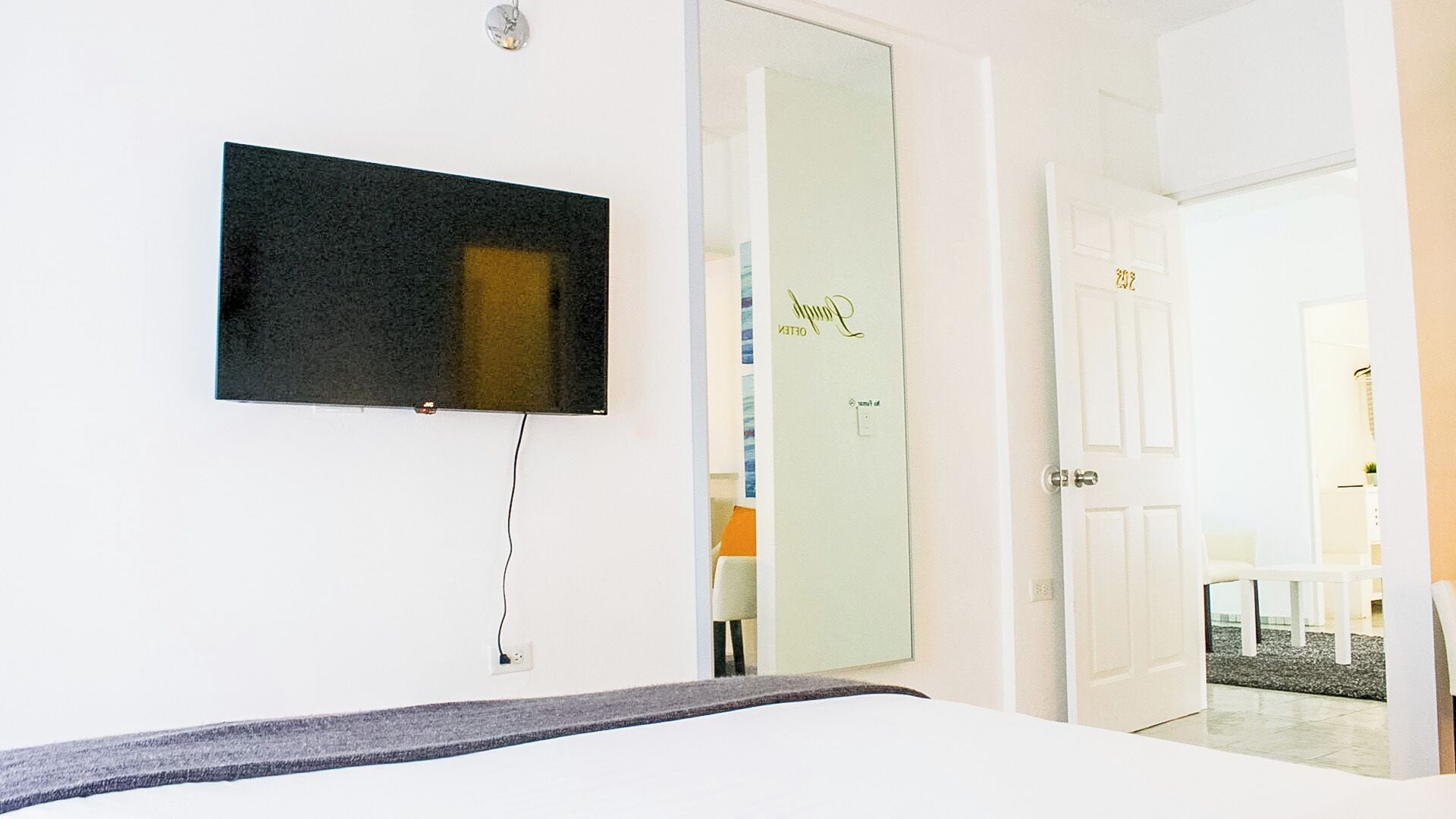 A hotel room with a bed, a flat-screen TV mounted on the wall, a door leading to a bathroom, and a small table with a chair.