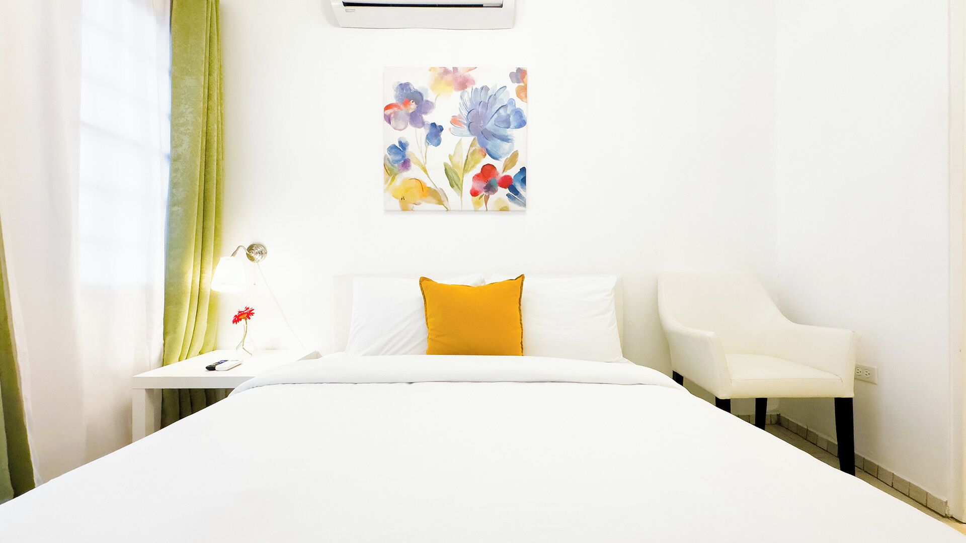 A hotel room with a bed, white walls, a painting of flowers, a nightstand with a lamp, and a chair.