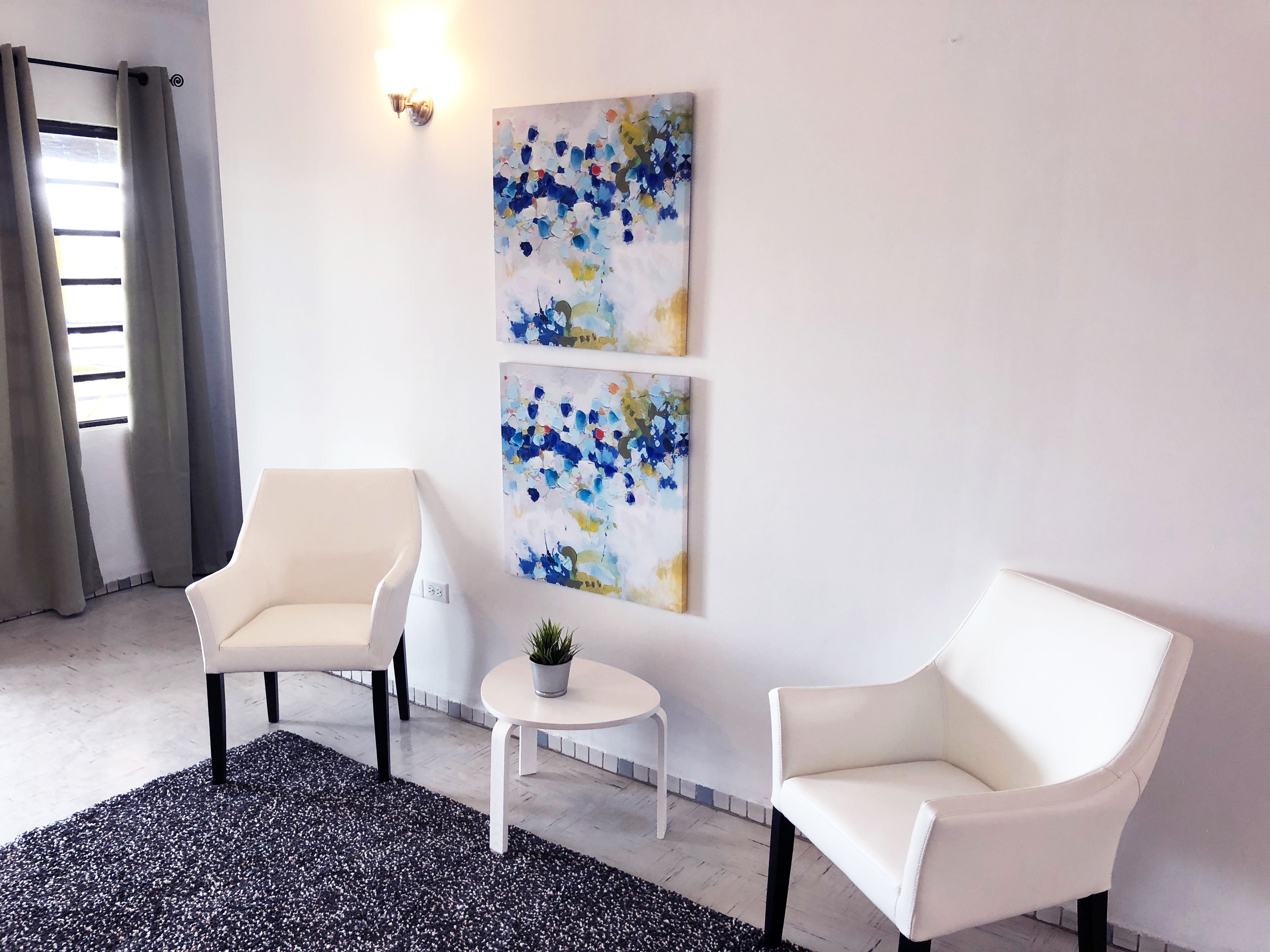 Ein modernes Zimmer mit zwei weißen Sesseln, einem kleinen runden Tisch mit Pflanze und abstrakter Kunst an der Wand.