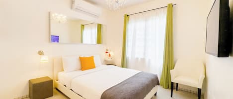 A hotel room with a bed, a chair, a desk with a lamp, a mirror, and a chandelier.